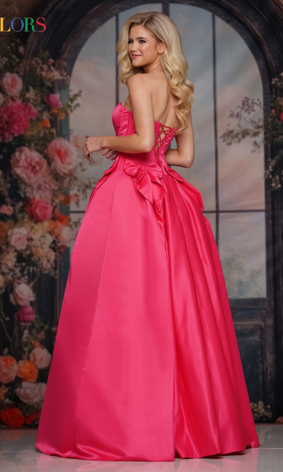 A woman in the Bow-Front Long Prom Ball Gown: Colors Dress 3739, with a vibrant pink strapless sweetheart neckline and lace-up back topped by a large bow, stands before a floral and arched window backdrop.